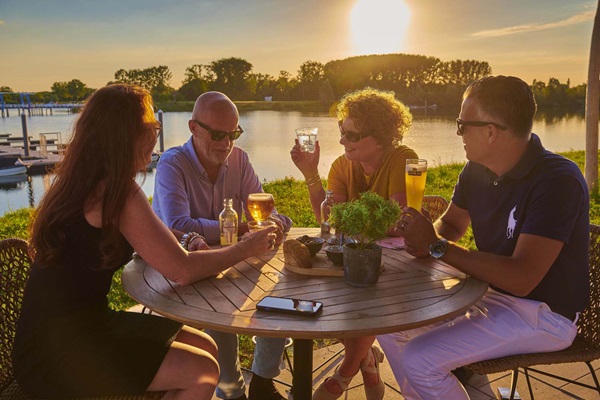 Twee vrouwen en mannen genieten van de ondergaande zon op het terras van Maas Residence Thorn