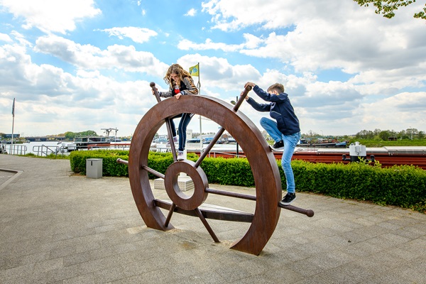Twee kinderen klimmen op een groot scheepsroer bij de haven, met boten op de achtergrond.