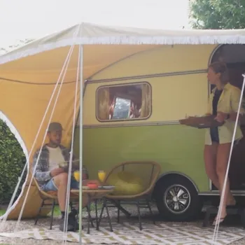 Stel bij een retrocaravan onder een luifel; man zit met ontbijt aan tafel, vrouw komt naar buiten met een dienblad.
