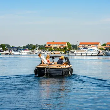 Een groepje mensen vaart in een kleine huurboot over het water, met jachthavens en vakantiehuisjes op de achtergrond. Ideaal voor een ontspannen dagje varen in Limburg.