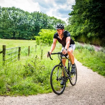 Gravelbiker fietst over een onverhard pad langs weilanden en bosrand op een zomerse dag.