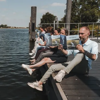 Mensen lezen het magazine Hartstocht aan de waterkant van de Maasplassen en genieten van het uitzicht over het water.