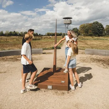Vier bezoekers bekijken een speervormig Archeo Route Limburg-kunstwerk op een archeologische locatie in het landschap.